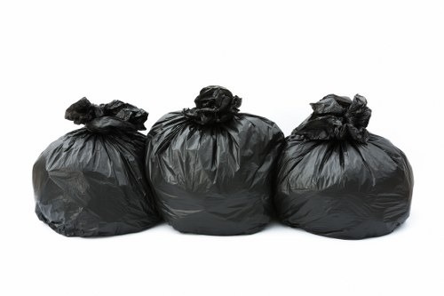 Three black plastic rubbish bags, tied at the top, are arranged in a row on a white surface, likely in preparation for waste collection or disposal. The bags appear full and are made of standard glossy, flexible plastic with visible creases and folds, indicating they are filled with waste material. They are positioned close together, with the middle bag slightly behind the others, suggesting a typical outdoor or commercial waste collection scene. The background is plain and white, emphasizing the bags without additional environmental context, consistent with imagery used by rubbish removal services such as Commercial Waste Norbiton. The image visually conveys the concept of waste management, suitable for illustrating waste collection or rubbish disposal services in the Norbiton area or similar urban locations in the UK.