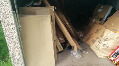 A collection of assorted rubbish and cardboard boxes stacked against a corner of a garage or storage area. The waste includes large, partially collapsed cardboard boxes, some flattened and others still in their original shape, with visible printed labels and branding. There is also a beige wooden or particleboard piece leaning against the wall, and a plastic water bottle on the floor nearby. The surrounding environment appears to be an outdoor space, possibly a driveway or side passage, with a concrete floor and a partial view of a textured wall and a grassy area outside. This scene represents typical waste materials that would require professional rubbish removal services, such as those offered by Commercial Waste Norbiton, operating in the Norbiton area, close to Kingston upon Thames, within the postal region postcodes around KT1.