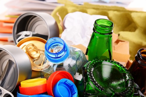 A detailed close-up image showing a collection of discarded glass bottles, plastic containers, and lids scattered randomly on a surface, likely in an outdoor or semi-indoor setting associated with waste disposal in Norbiton. The green glass bottles are prominent, with some lying on their sides, their textures smooth and glossy, reflecting light. Transparent plastic bottles with blue caps and various plastic lids in red, yellow, and blue colors are visible among the debris, indicating mixed waste typical of household or commercial refuse. In the background, there are blurred cardboard and paper materials, suggesting a cluttered environment ready for rubbish collection or removal. The image visually communicates the type of waste managed by commercial waste removal services like Commercial Waste Norbiton, emphasizing the importance of responsible disposal of glass and plastic waste within the local postcode area, aligning naturally with the services offered in Norbiton and surrounding districts.