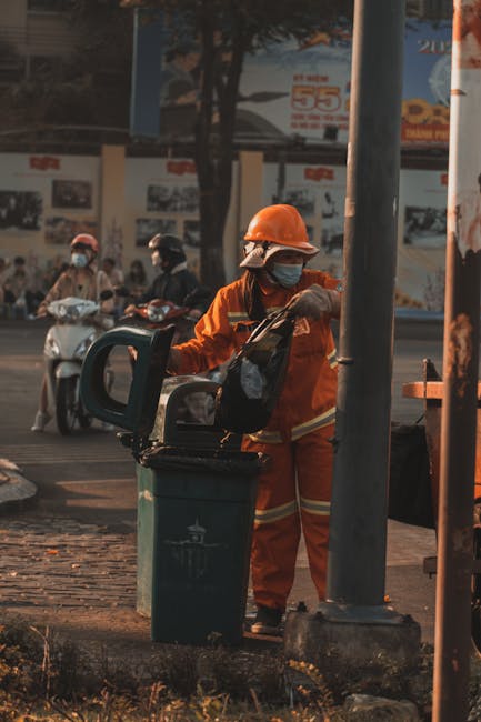 A worker dressed in bright orange high-visibility clothing and a safety helmet is using a hand-held tool to lift or empty a green waste collection bin positioned on a paved sidewalk. The worker is wea