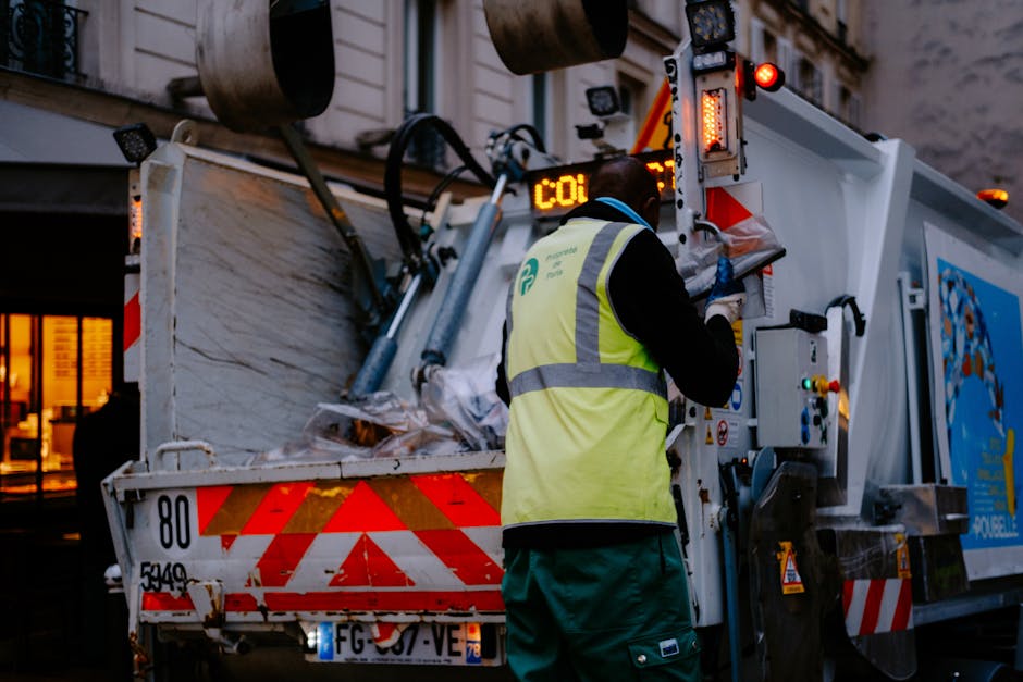 A worker dressed in bright orange high-visibility clothing and a safety helmet is using a hand-held tool to lift or empty a green waste collection bin positioned on a paved sidewalk. The worker is wea