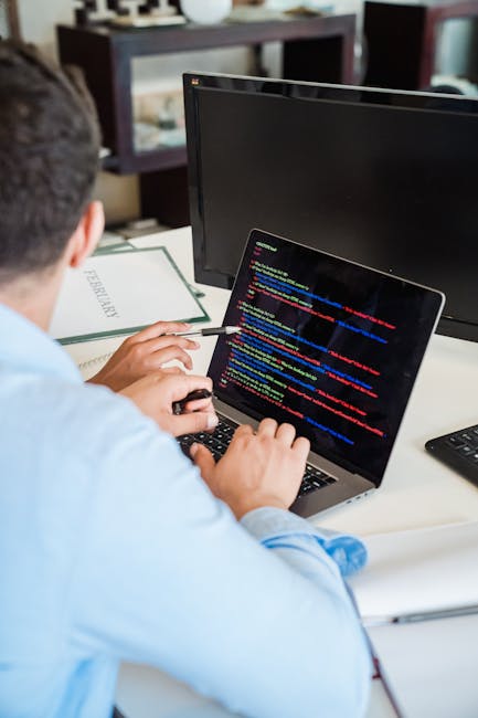 A man with dark hair, glasses, and a beard is seated at a wooden desk, working on a computer with dual monitors. The left monitor displays a file management interface with a dark theme, showing numero
