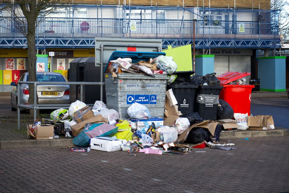A man dressed in a black jacket, grey beanie, and white gloves is standing on a train station platform beside a red wheeled trolley loaded with numerous large garbage bags in black, grey, and orange, 